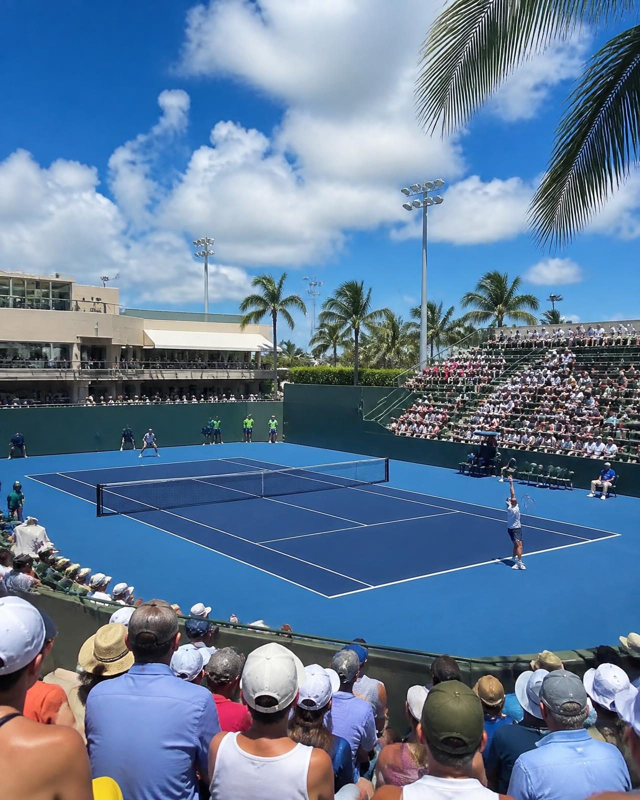 An outdoor blue hard court surrounded by palm trees under a bright sky