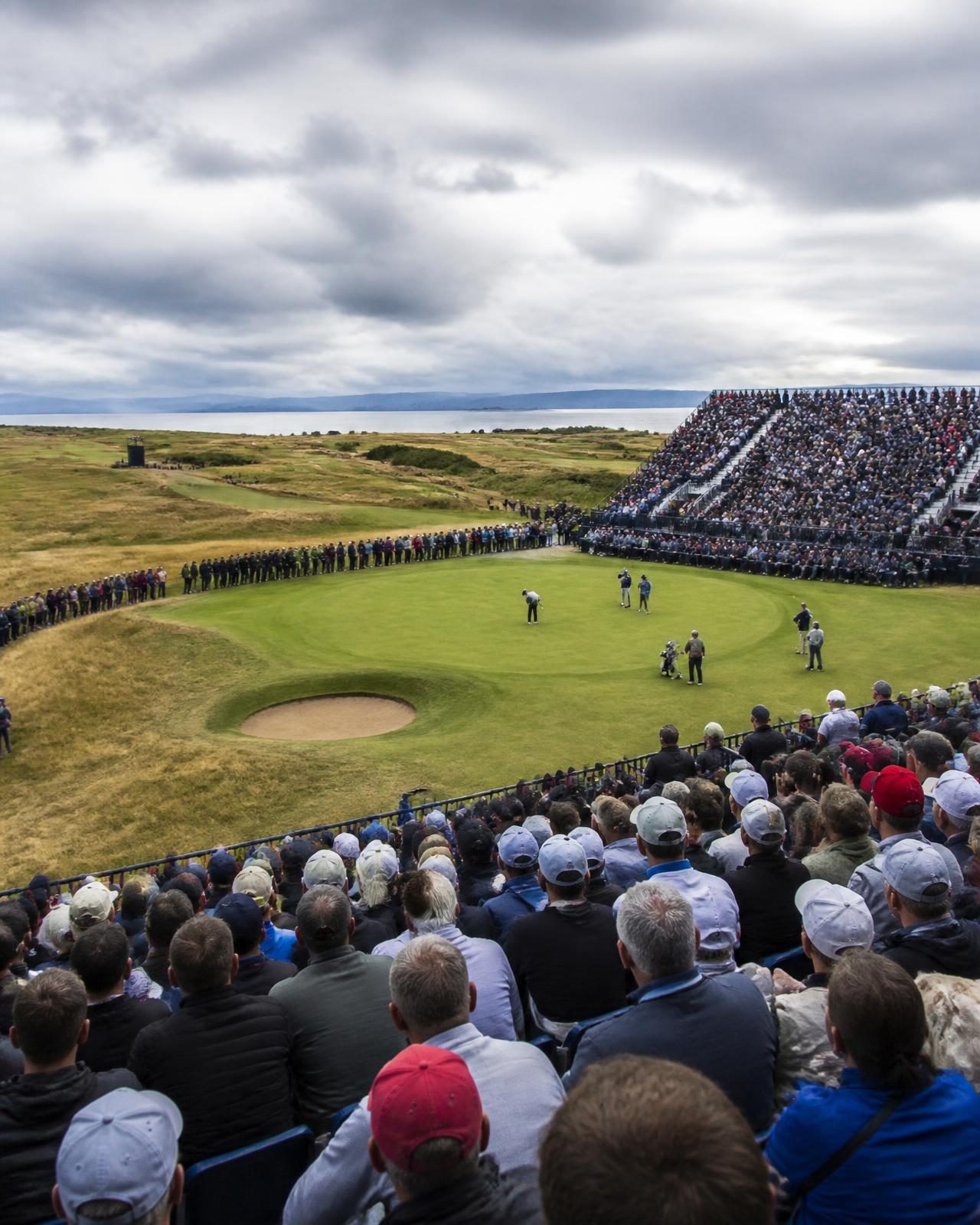 A packed grandstand around a links golf green under dramatic clouds
