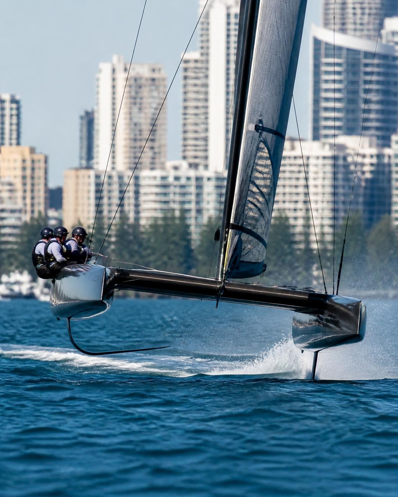 A foiling racing catamaran at speed in front of a waterfront city skyline