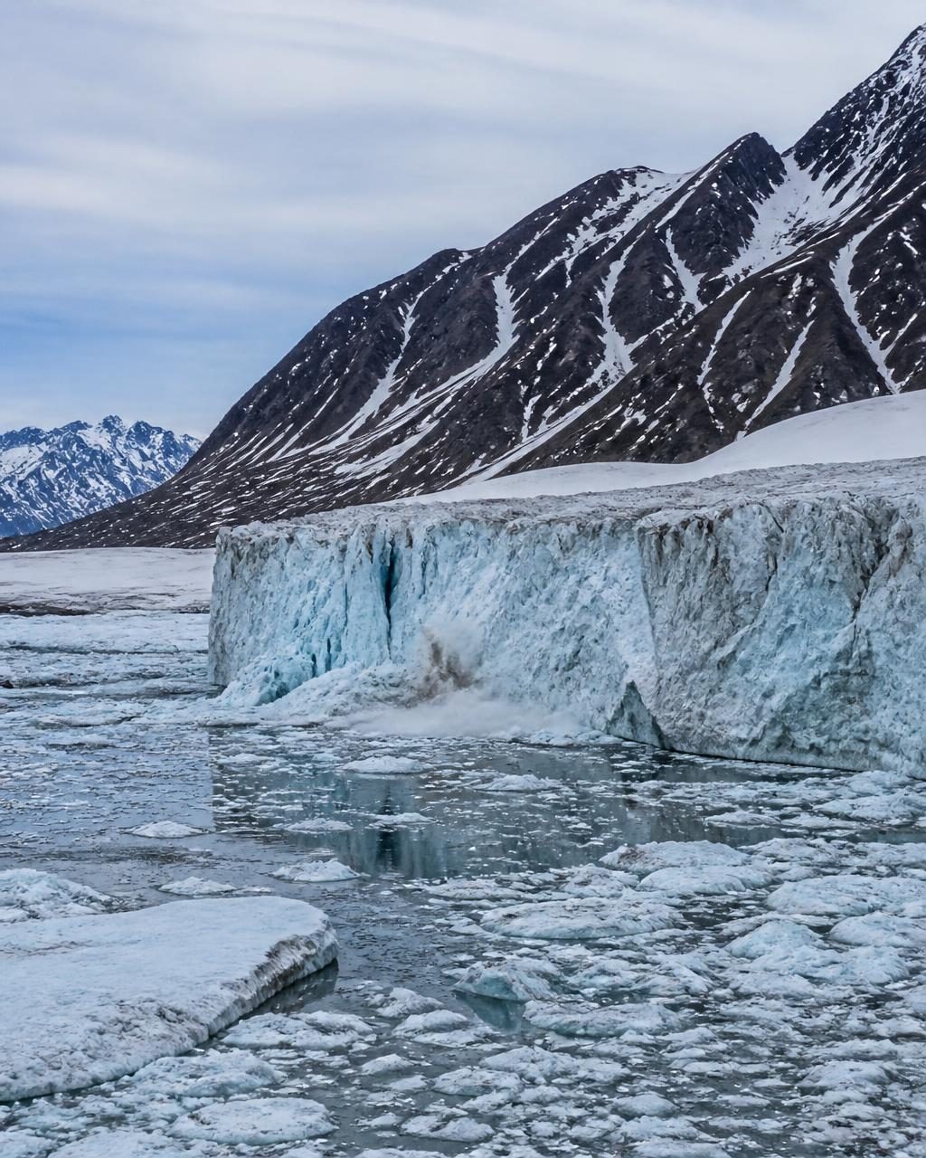 Glacier front calving into ice-strewn arctic water below snow-streaked mountains