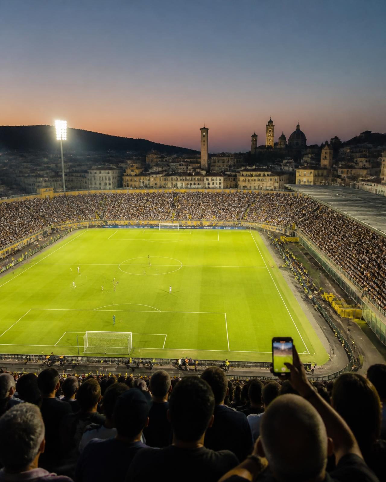 A European football stadium at dusk with a historic city skyline behind